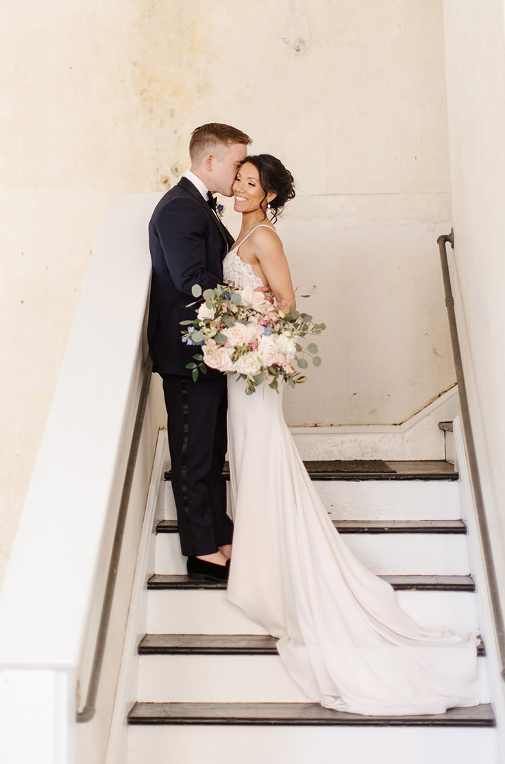 Groom embraces his bride on stairs and in a lace bodice wedding dress with a crepe fit and flare skirt, holding a bouquet. 