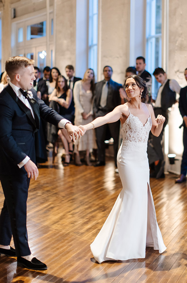 Groom dances with bride in a V-neck lace bodice gown with a crepe fitted skirt and skirt slit, in a ballroom near guests.