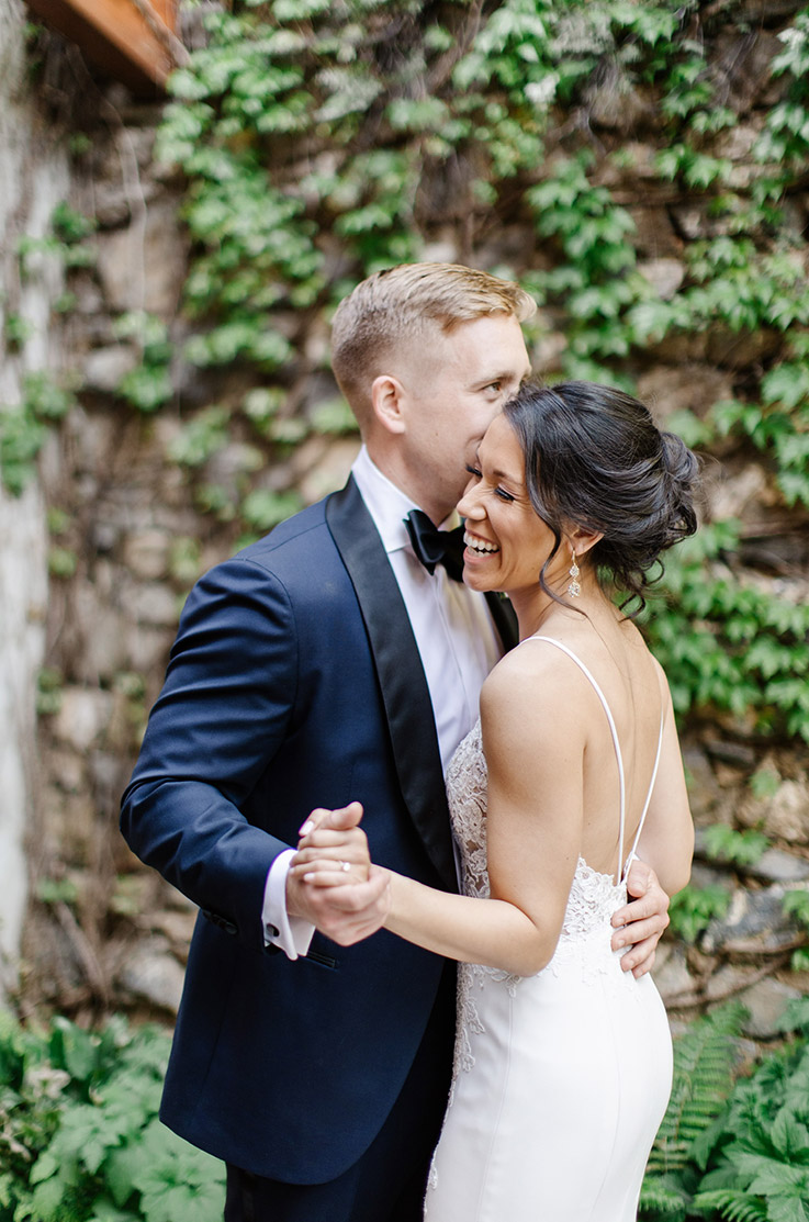 Bride in a low back lace gown with a crepe fit and flare skirt and thin straps, is dancing with groom by an ivy-covered wall.