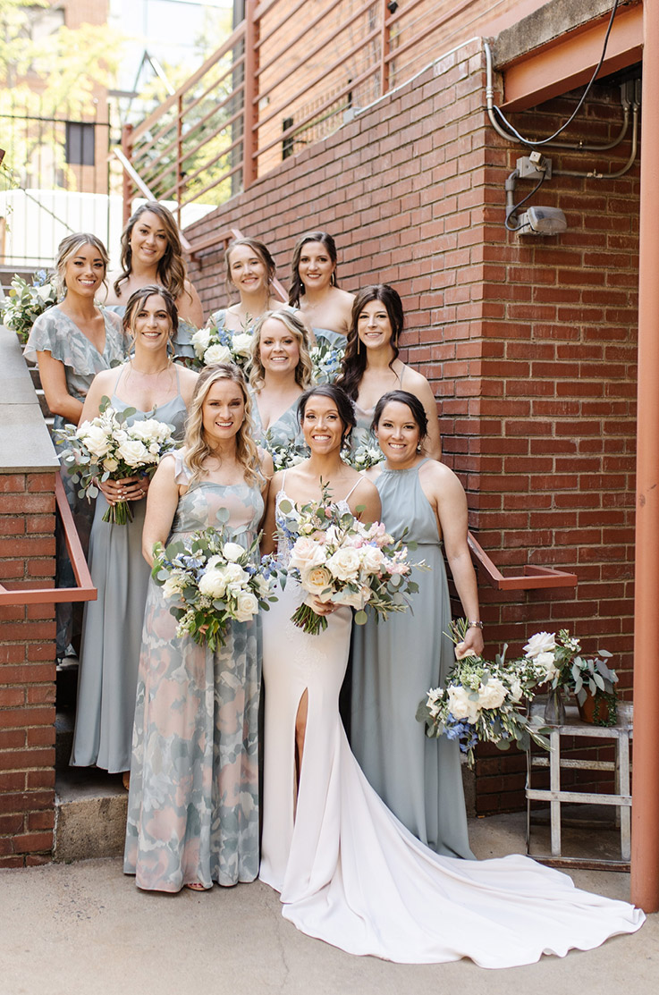 Bride in a crepe fit and flare gown with a skirt slit, poses with bridesmaids holding bouquets at a brick building.