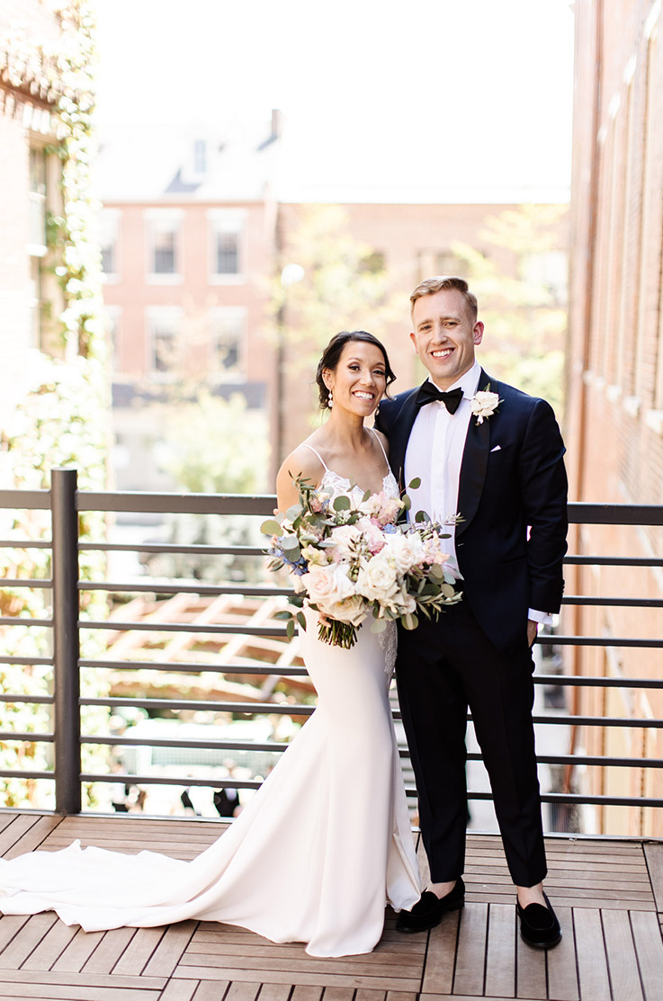Bride in a V-neck lace bodice gown with fit and flare skirt, poses next to groom by a railing on an outdoor terrace.