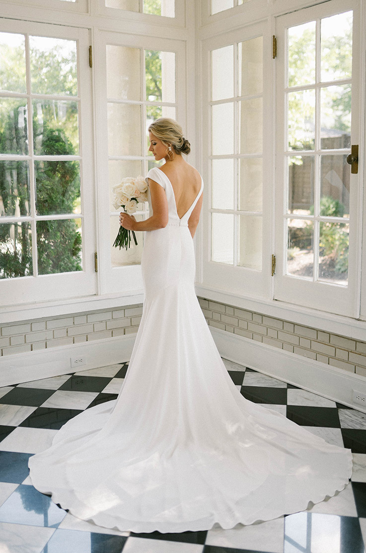 Bride poses in a crepe fit and flare gown with a low open back, holding a white bouquet near on a checkered floor in sunroom.