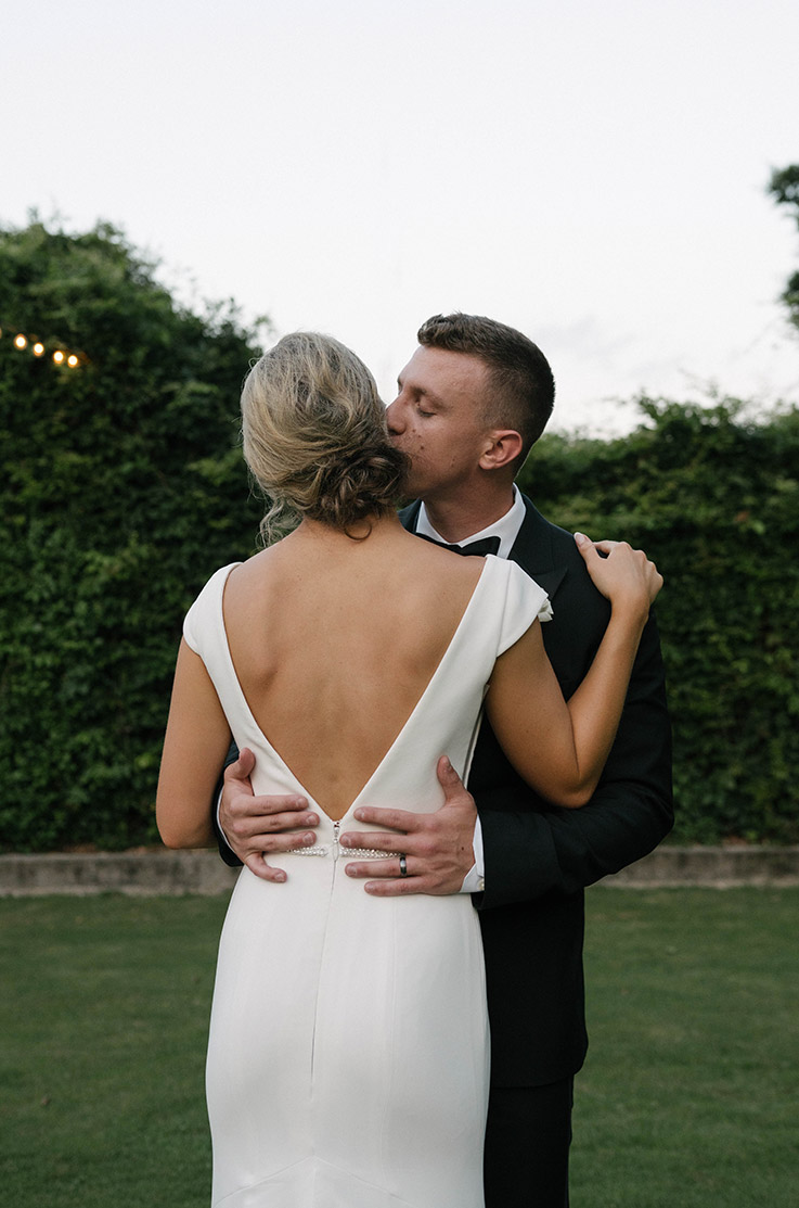 Groom embraces bride in a deep-V back crepe wedding dress with cap sleeves, standing on an open green lawn as the sun sets. 