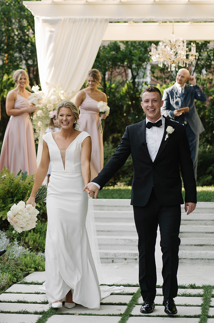 Bride in a plunging neck crepe gown with a fit and flare skirt, veil and a belt, walks hand in hand with groom in the aisle.