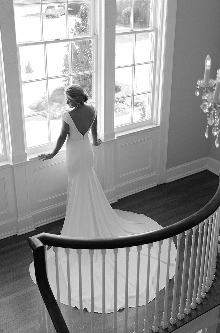 Bride faces windows and leans on windowsill in a V-back fit and flare crepe gown with cap sleeves by a railing and chandelier. 