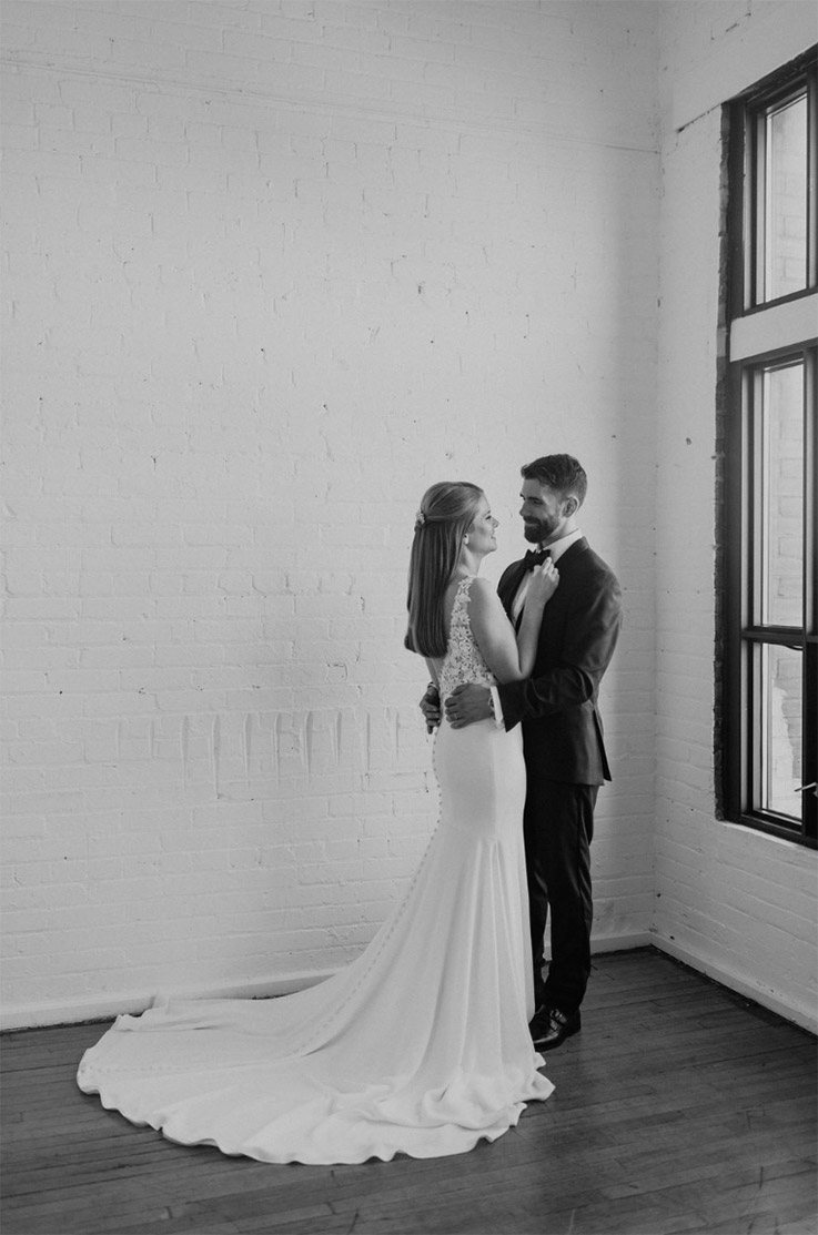 Groom embraces bride in a crepe fit and flare wedding dress with buttons and a lace V-back, indoors in black and white.