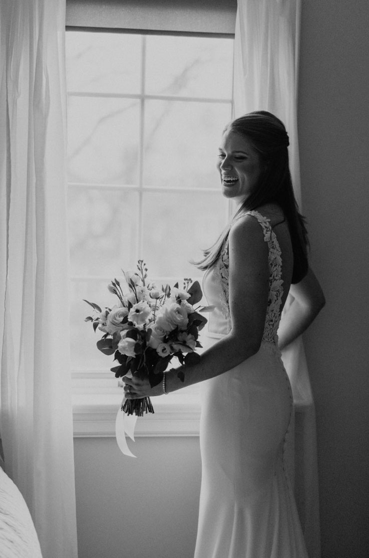 Bride stands by a window, in a crepe fit and flare gown with a lace back and sides, holding a bouquet in a brightly lit room. 