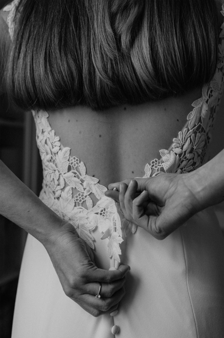 Close-up of bride zipping up into a V-shaped back lace gown with a crepe skirt and button detail, in black and white.