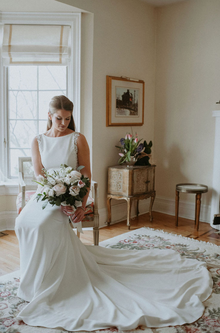 Bride poses sitting in a chair, in a boat neck crepe gown with a flowing train and lace details, holding a bouquet indoors.