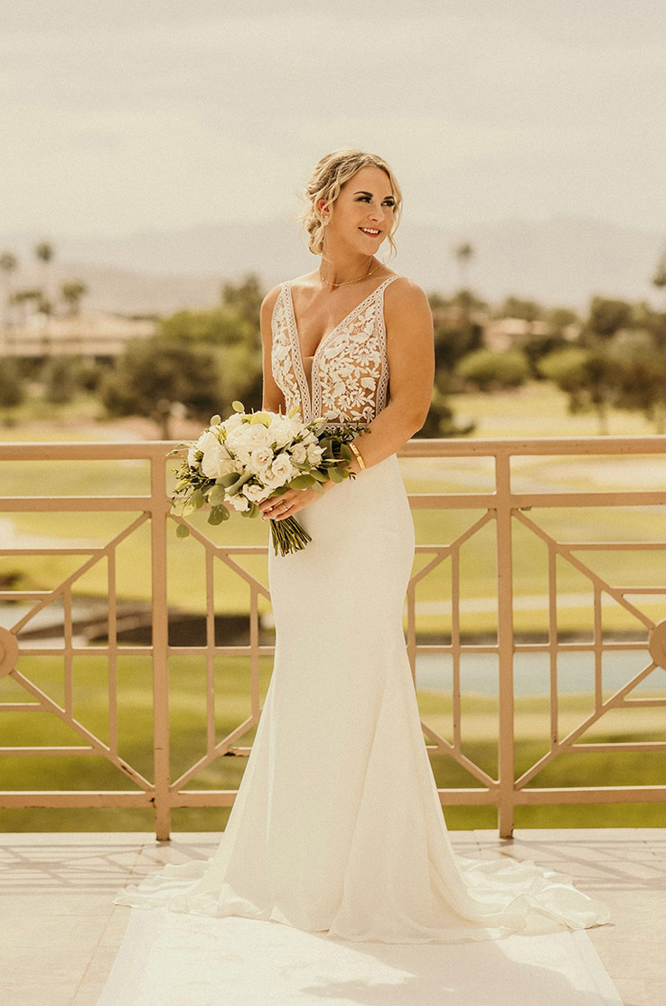 Bride poses on a terrace in a lace V-neck gown with side cutouts and a crepe fit and flare skirt, is holding a bouquet. 