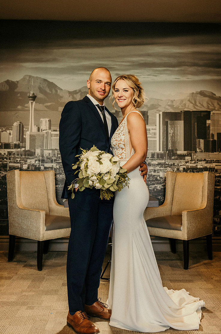 Bride in a lace bodice wedding dress with a fit and flare crepe skirt, holds a bouquet beside groom at a city skyline backdrop.