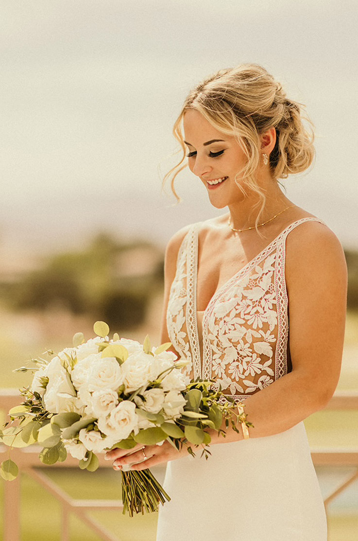 Close-up of bride in a sleeveless lace bodice gown with a V-neck, cutouts and a crepe fitted skirt, holding a white bouquet.