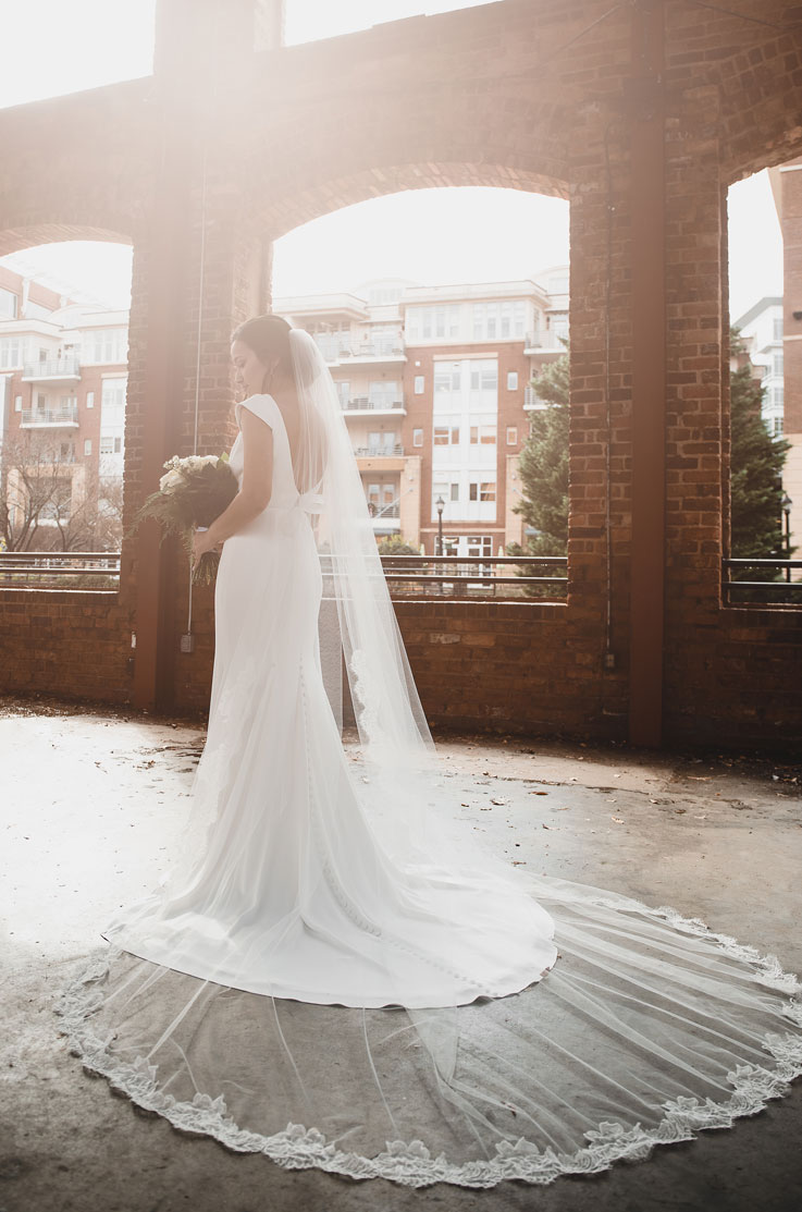 A bride in a cap sleeve wedding dress and veil, holding a bouquet, stands in an industrial brick building with her dress train and veil fanned out behind her.