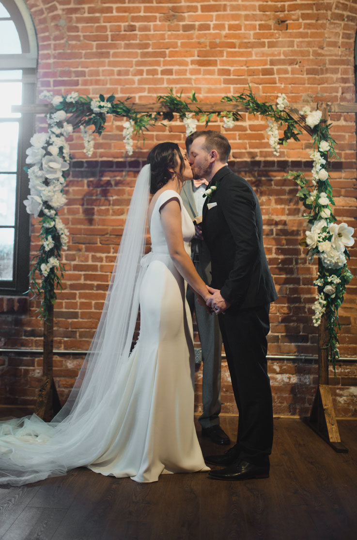 A bride in a cap sleeve wedding dress and veil, and a groom in a suit hold hands and kiss in front of a man in a suit and a floral ceremony arch, inside a brick building.