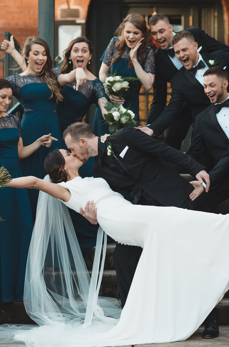 A groom in a suit dips a bride in a cap sleeve wedding dress and veil as he kisses her, with their bridal party cheering around them.