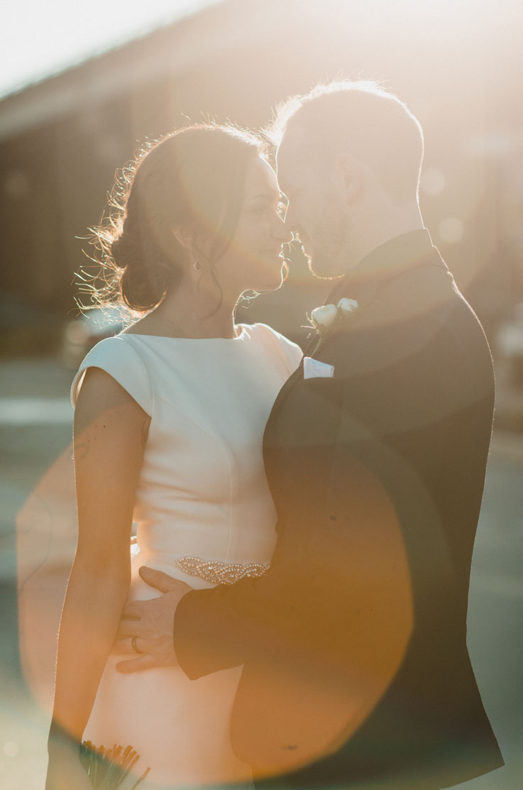 A bride in a cap sleeve wedding dress and a groom in a suit hold each other, about to kiss, outside with the sun shining behind them.