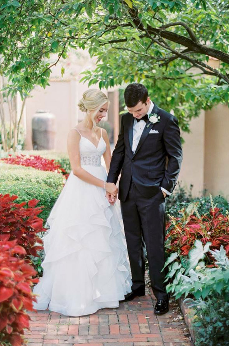 A bride in a flowing tulle wedding dress and a groom in a suit stand outside in a garden, on a brick path and tree branches above them, as they look down and smile.