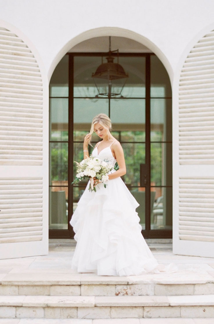 A bride in a flowing tulle wedding dress, holding a bouquet, poses in front of arched double slatted doors at the top of a set of stairs, with a large window behind her.