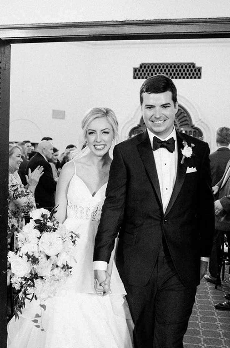 A bride in a flowing tulle wedding dress, holding a bouquet, and a groom in a suit, hold hands and walk through an entrance with wedding guests standing behind them.