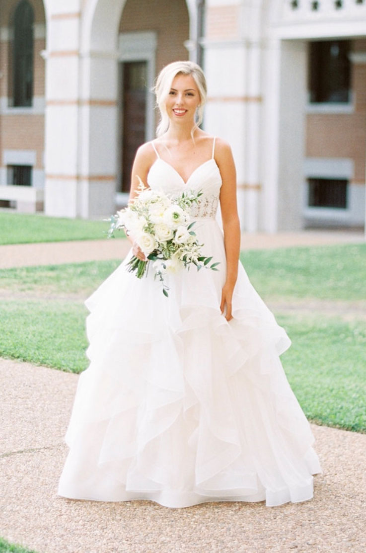 A smiling bride in a flowing tulle wedding dress, holding a bouquet, poses outside on a path outdoors with a grass yard and a building behind her.