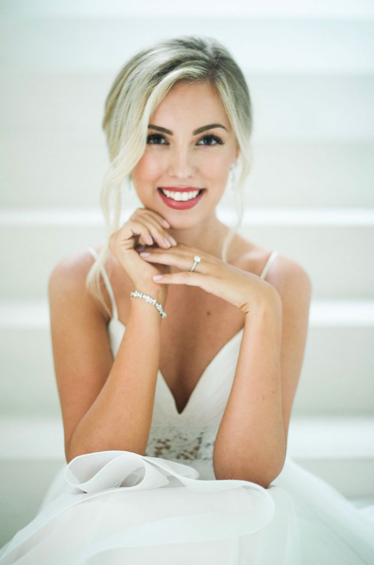 A bride in a V-neck wedding dress sits on a set of stairs indoors and smiles, with her elbows on her knees and her hands up close to her chin.