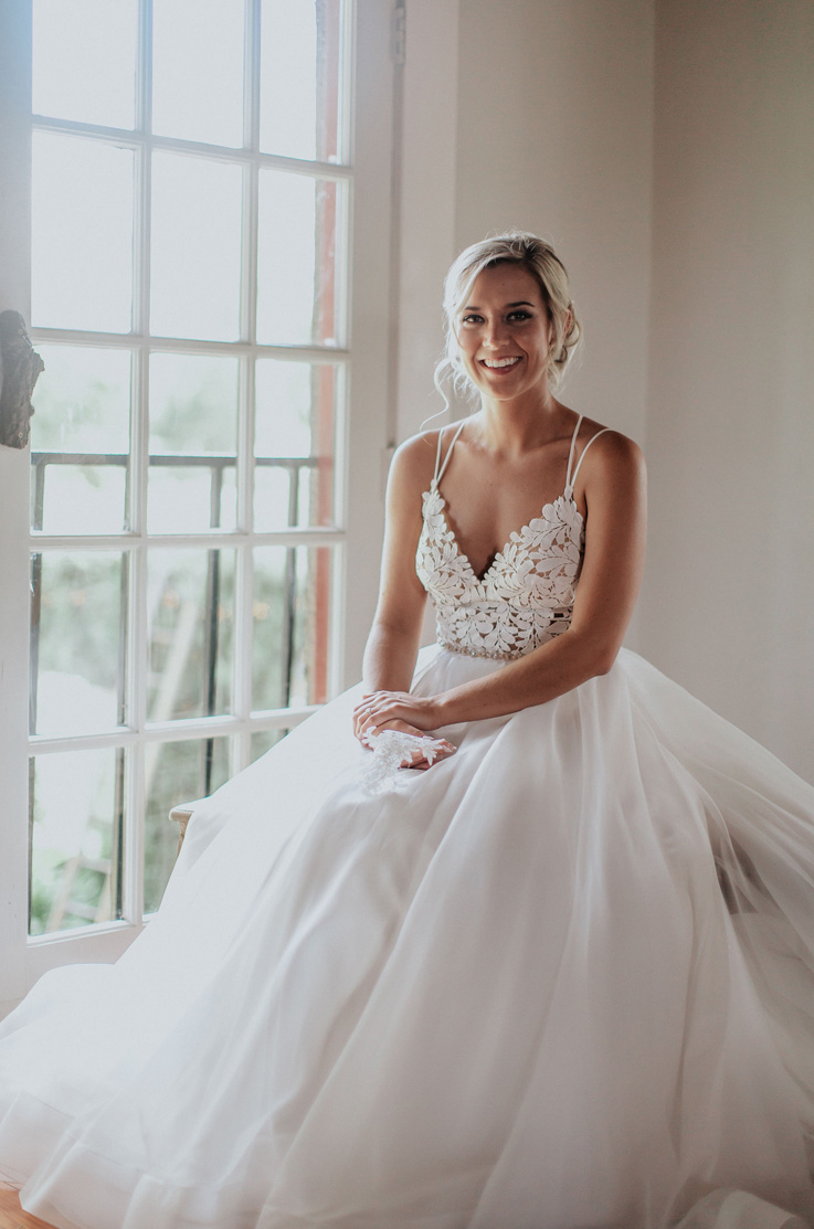 A bride in a V-neck wedding dress sits indoors beside a window, with her hands folded in her lap as she smiles.