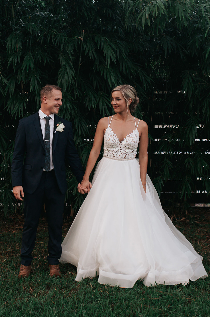 A bride in a V-neck wedding dress and a groom in a suit hold hands and look at each other, while standing outdoors in a grass yard with trees and a wooden fence behind them.