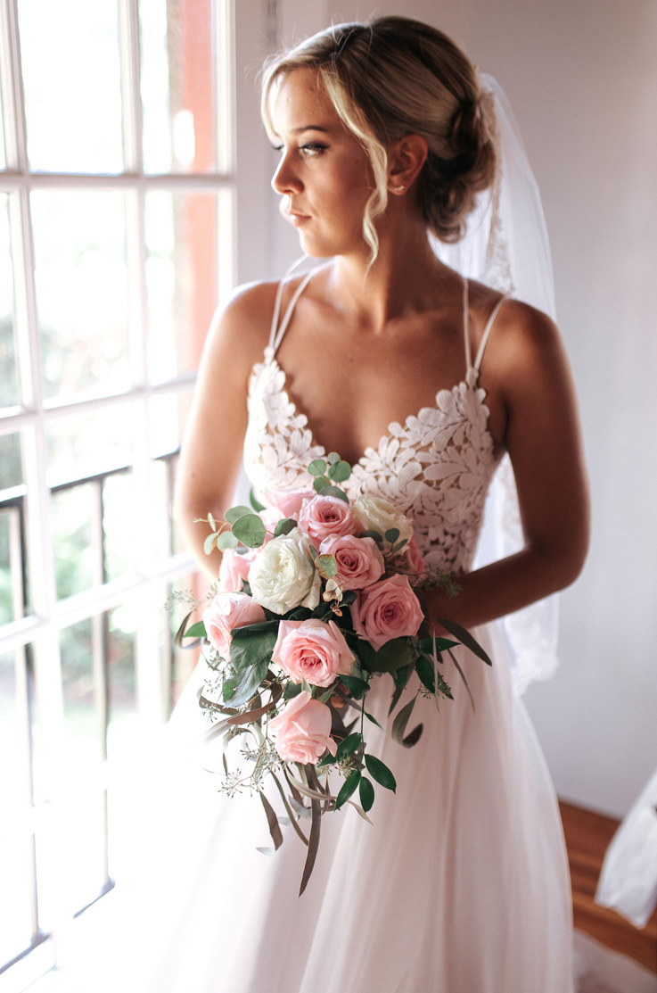 A bride in a V-neck wedding dress and veil, holding a bouquet, stands indoors beside a window as she looks to the side, towards the window.