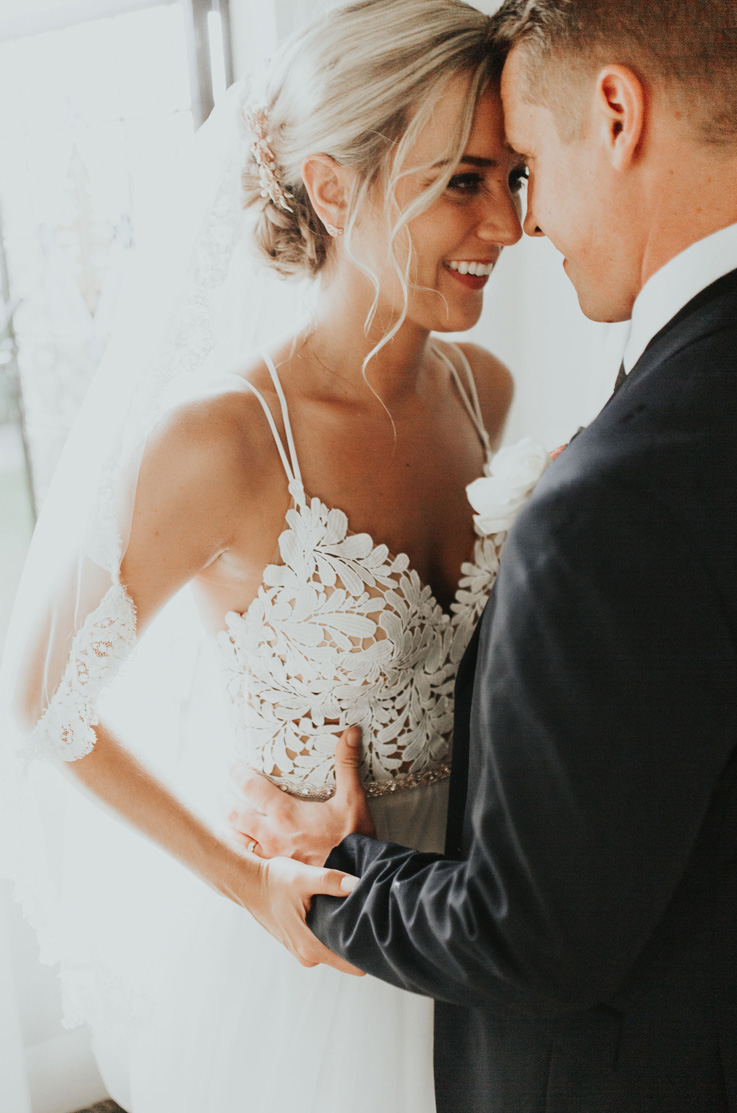 A bride in a V-neck wedding dress and veil, and a groom in a suit stand close together facing each other and smile, with a brightly lit background.