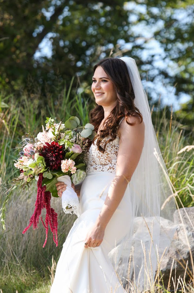 A bride in a fitted wedding dress and veil, holding a bouquet, looks into the distance and smiles as she stands outdoors with tall grass and trees behind her.