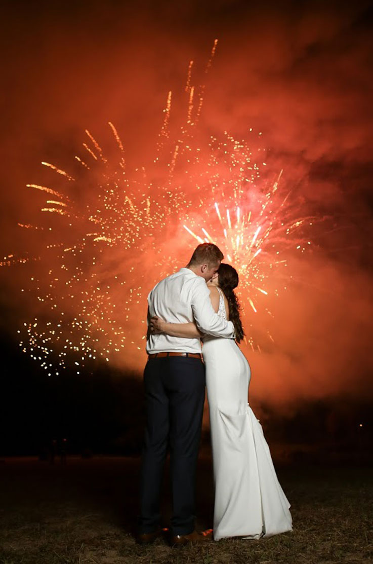A bride in a fitted wedding dress and a groom in a dress shirt hold each other and kiss outside at night, with fireworks going off in the sky behind them.