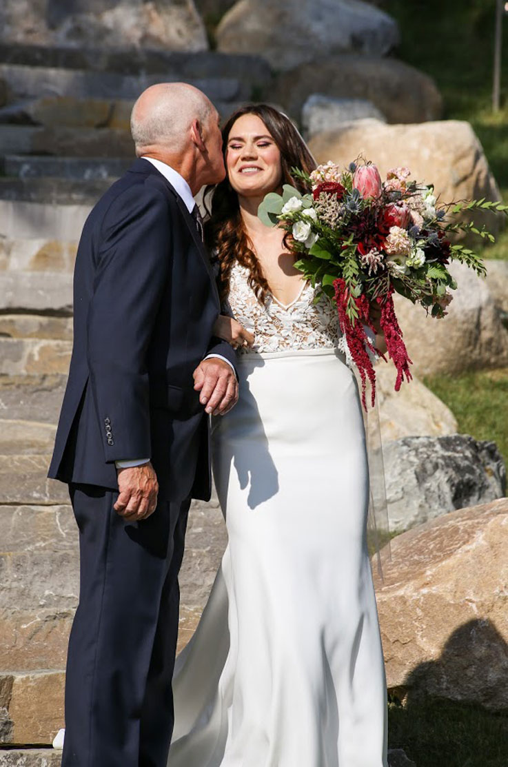 A bride in a fitted wedding dress, holding a bouquet, gets a kiss on the cheek from a man in a suit as they stand outside in front of set of rock stairs.