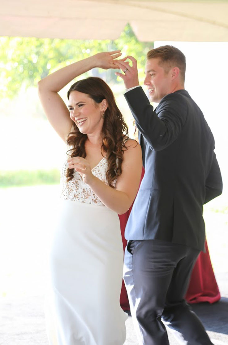 A bride in a fitted wedding dress and a groom in a suit dance in an outdoor tent as they smile.