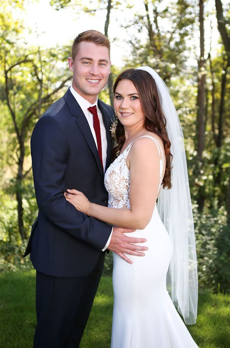 A bride in a fitted wedding dress and veil and a groom in a suit smile and hold each other while standing outside with trees in the background.