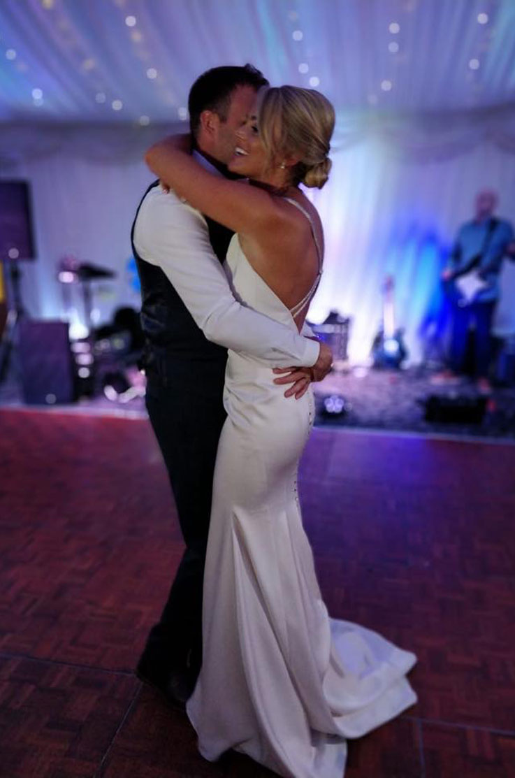 A smiling bride in an embellished wedding dress and a groom in a suit jacket hold each other close on a dancefloor, with a band and sparkling lights behind them.