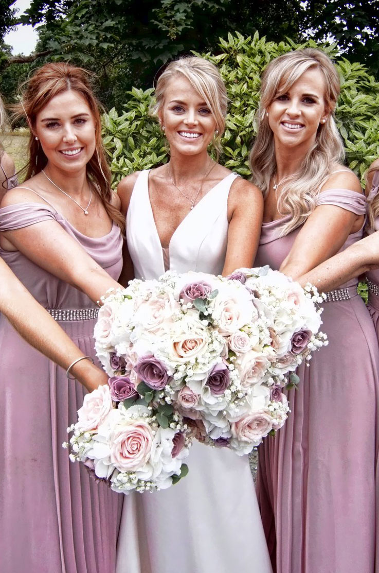 A bride in a plunging neckline wedding dress stands outside with a few bridesmaids, all of them smiling and holding their bouquets together with trees and bushes in the background.