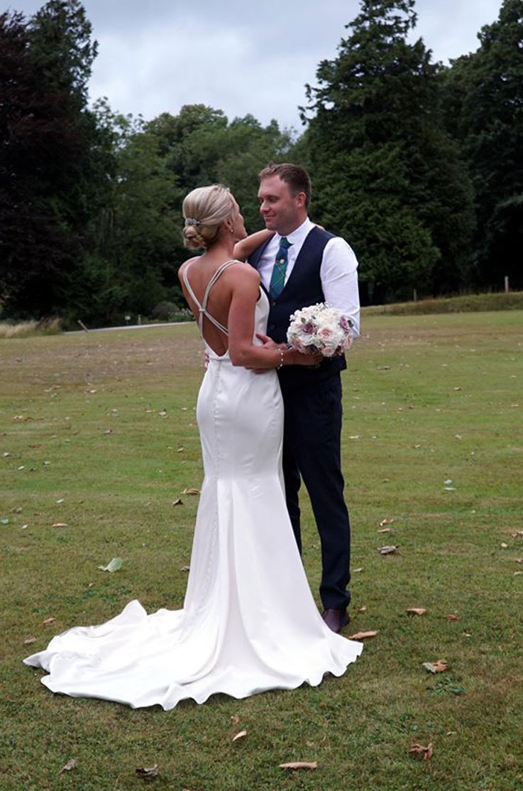 A bride in an embellished wedding dress, holding a bouquet, and a groom in a suit hold each other while facing each other, outside in a grass field, with trees behind them.