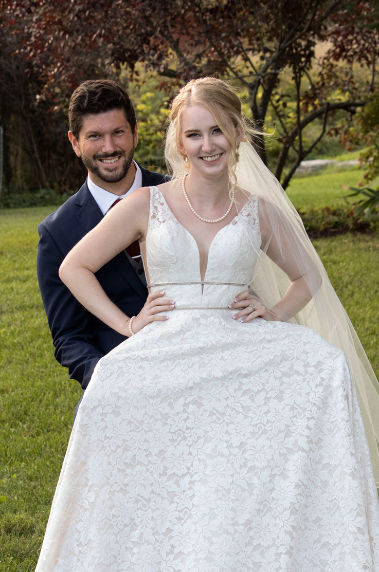A bride in an embellished lace wedding dress and veil, and a groom in a suit smile and pose outside on a grass lawn, with trees behind them.
