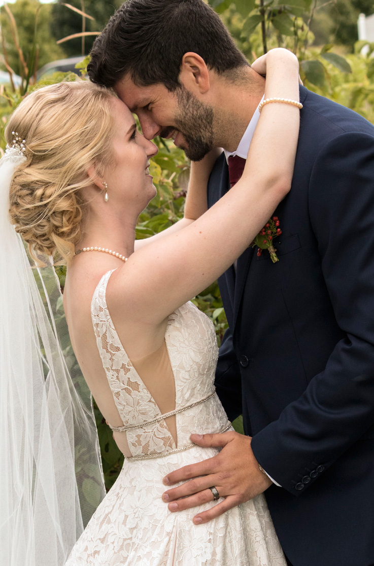 A bride in an embellished lace wedding dress and veil, and a groom in a suit, stand outside together facing each other and smiling, with their arms around each other.