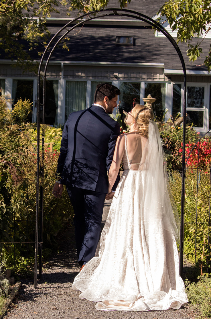 A bride in an embellished lace wedding dress and a groom in a suit hold hands and walk under an iron arch into a garden outdoors, with a house behind them.