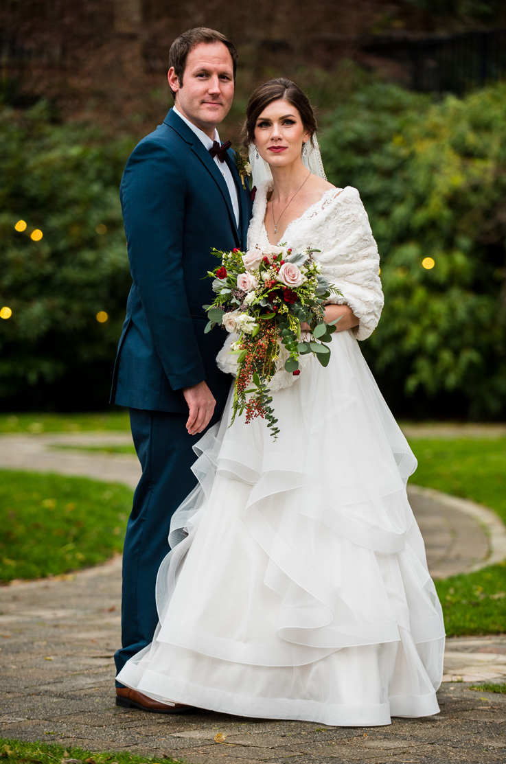 A bride in a tulle wedding dress and fur shawl, holding a bouquet, and a groom in a suit, stand together on a brick path outside, with trees in the background.