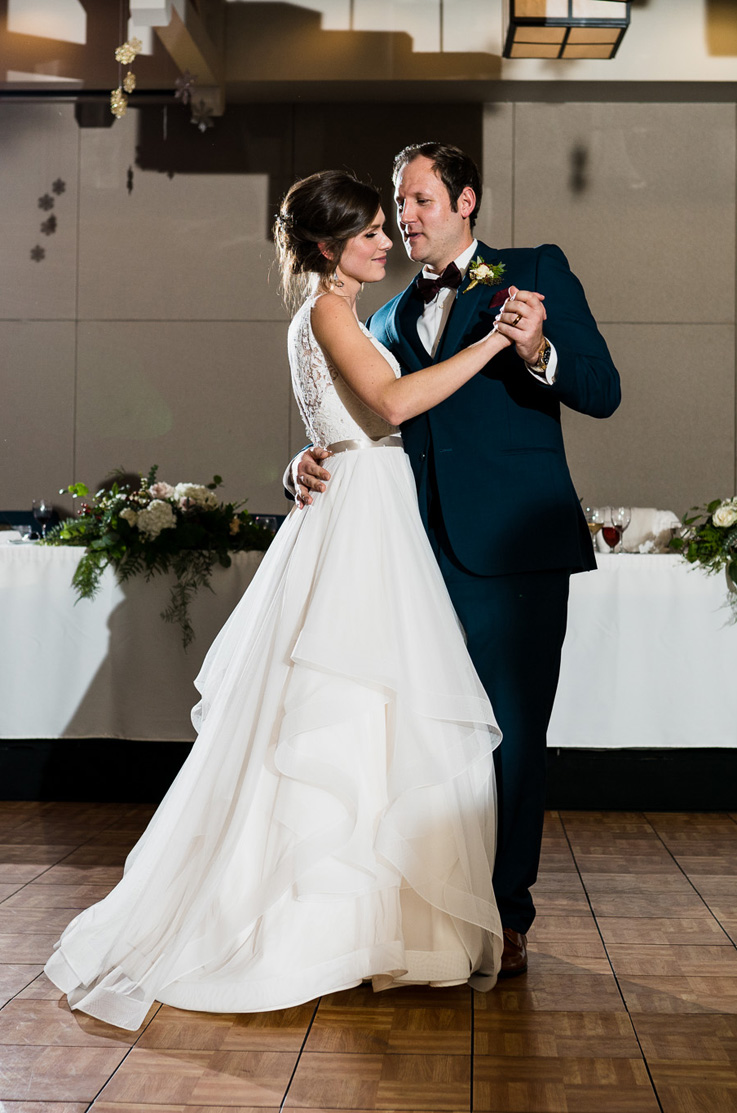 A bride in a beaded wedding dress and a groom in a suit dance on an indoor wooden dance floor, in front of the head table at a reception.