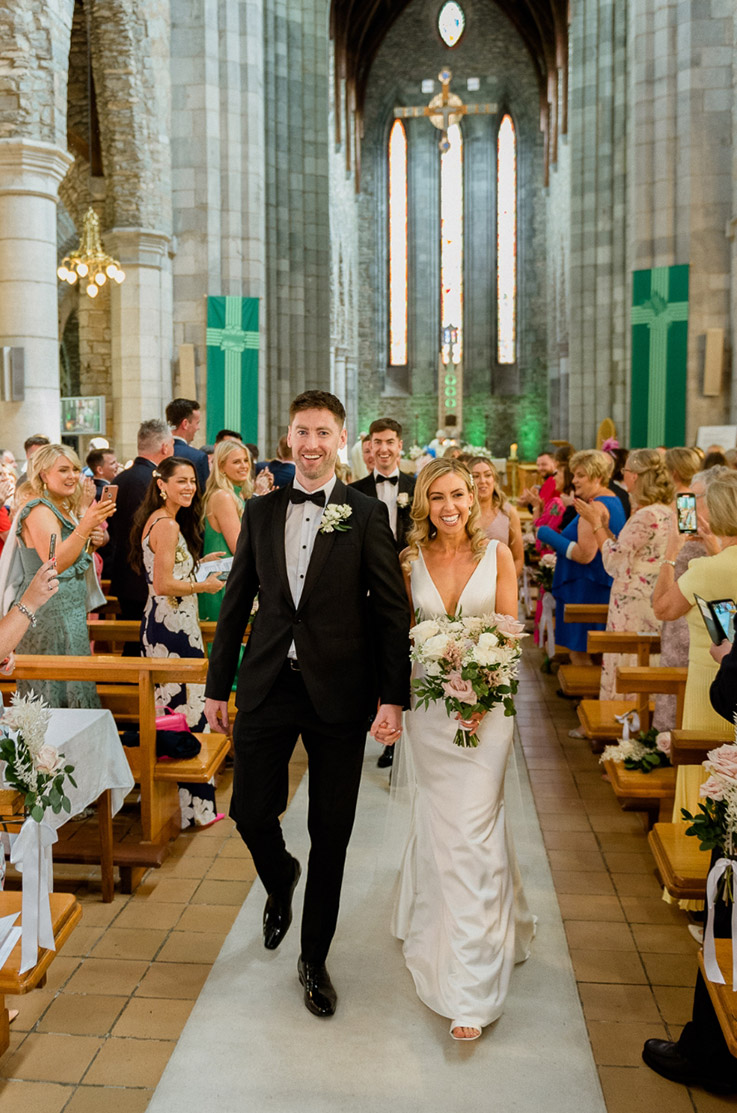 Bride in a plunging neck gown with a fit and flare skirt, holds a bouquet and walks alongside groom in the aisle in a chapel.