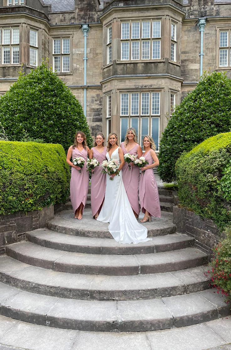 Bride in a satin square neck gown with fit and flare skirt, stands next to bridesmaids holding bouquets atop of stone steps. 