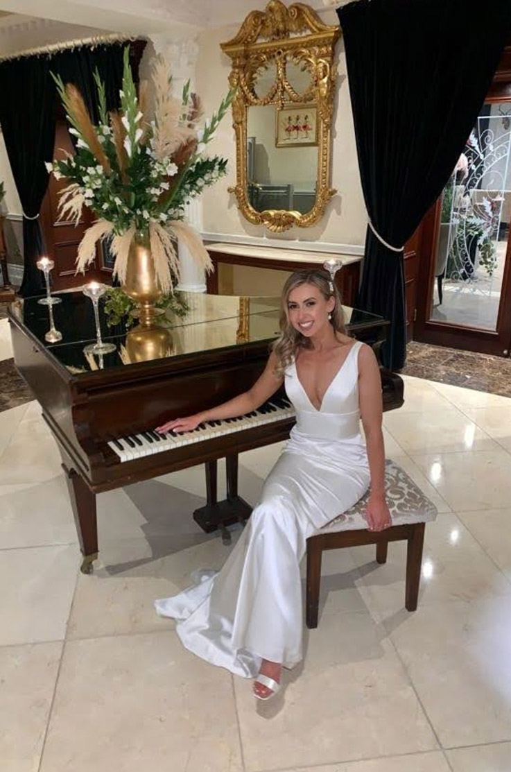 Bride in a sleeveless satin gown with a plunging neckline and an empire waist, sits at a grand piano in an elegant venue.