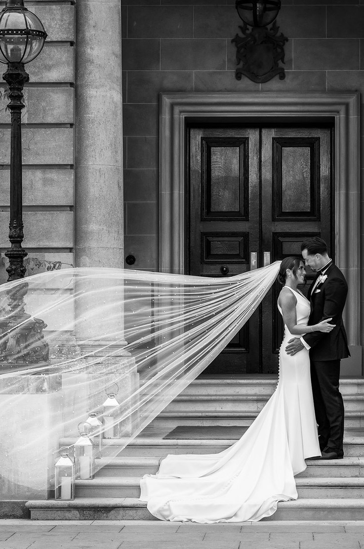 Groom embraces bride in a sleek crepe gown with buttons and a pearl edge veil standing on stairs in black and white.
