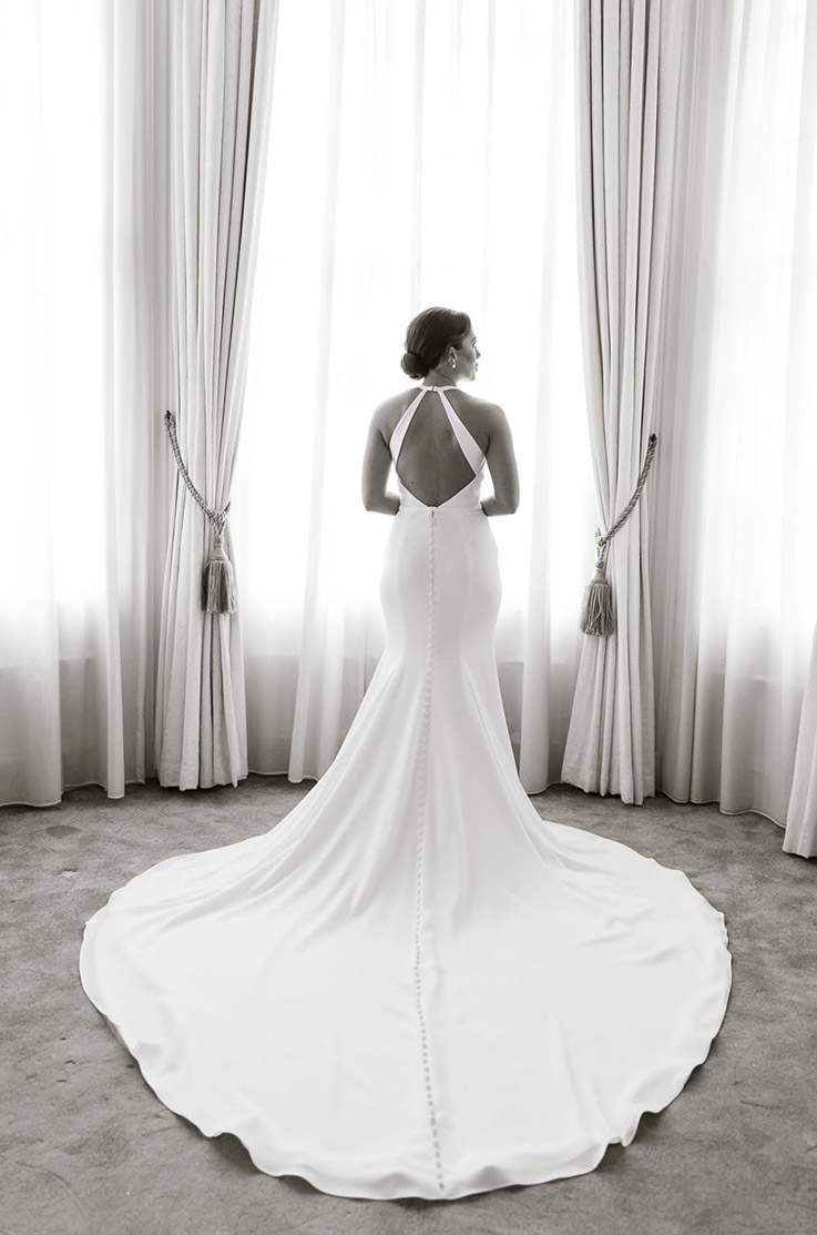 Bride faces a window in a brightly lit room, in a crepe fit and flare gown with a keyhole back and buttons down to the train.