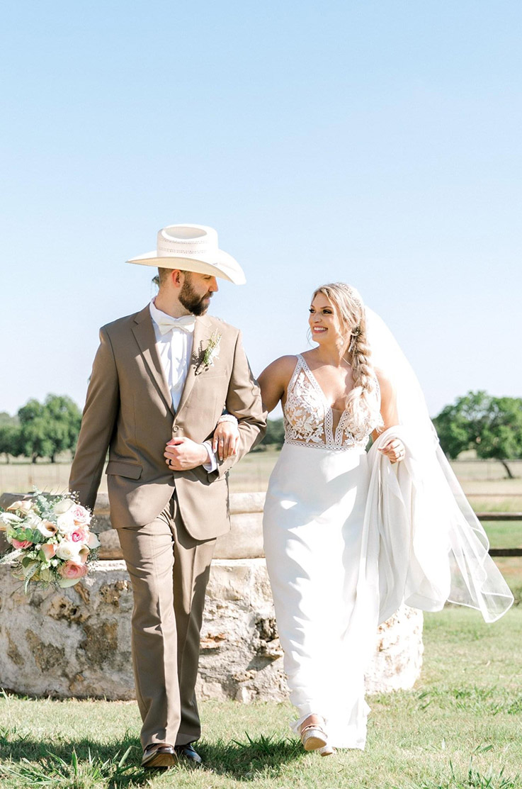 Groom walks with his bride in a lace bodice gown with a crepe A-line skirt and a veil, is holding her train in a sunny open ranch.