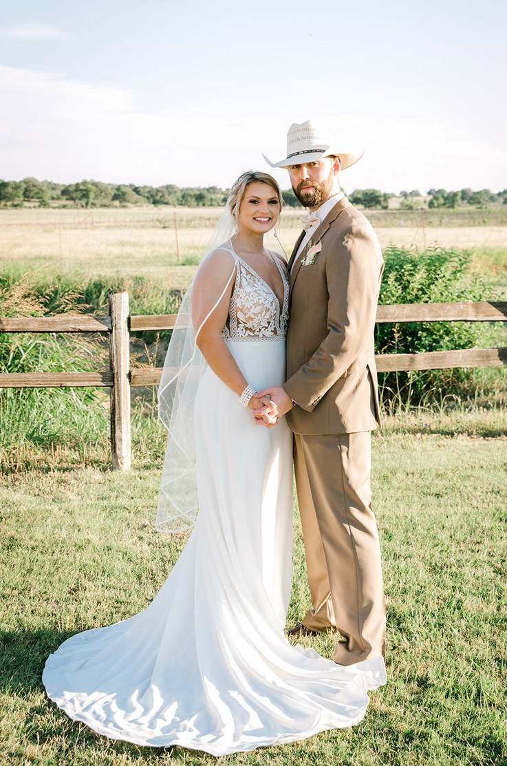 Bride in a V-neck lace wedding dress with a crepe A-line skirt and a veil, poses with groom on a countryside landscape.