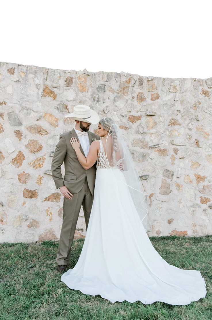 Bride in a deep-V back lace gown with an A-line crepe skirt and a veil, is posing with the groom by a stone wall outdoors.